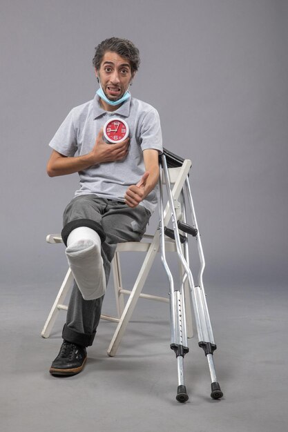 Vorderansicht junger Mann mit gebrochenem Fuß und Verband mit roter Uhr auf grauer Wand Twist Bein menschlicher Schmerz männlicher Fuß