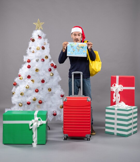 Vorderansicht junger Mann mit gelbem Rucksack, der Karte mit beiden Händen in der Nähe des weißen Weihnachtsbaums auf grauem isoliert hält