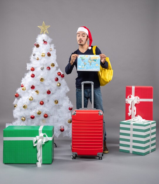 Vorderansicht junger Mann mit gelbem Rucksack, der Karte mit beiden Händen in der Nähe des weißen Weihnachtsbaums auf grauem isoliert hält