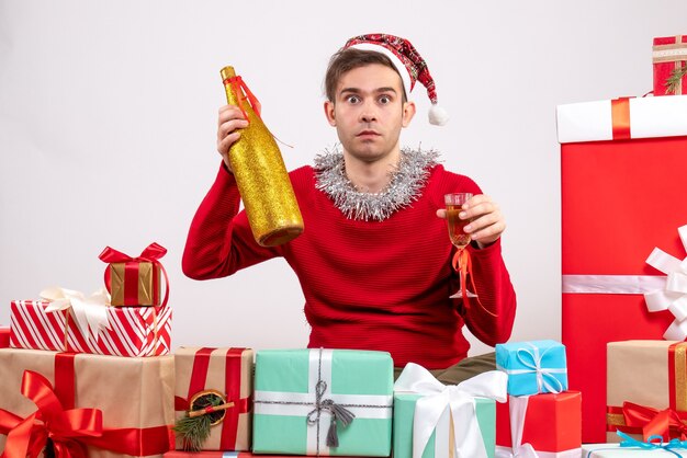 Vorderansicht-junger Mann mit großen Augen und Weihnachtsmütze, der Champagner hält, der um Weihnachtsgeschenke sitzt