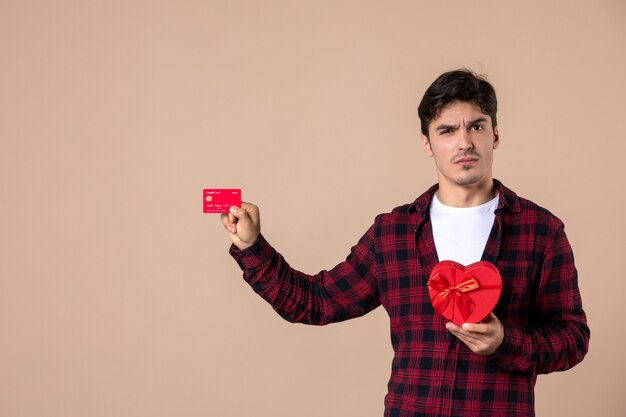 Vorderansicht junger Mann mit herzförmigem Geschenk und Bankkarte an brauner Wand