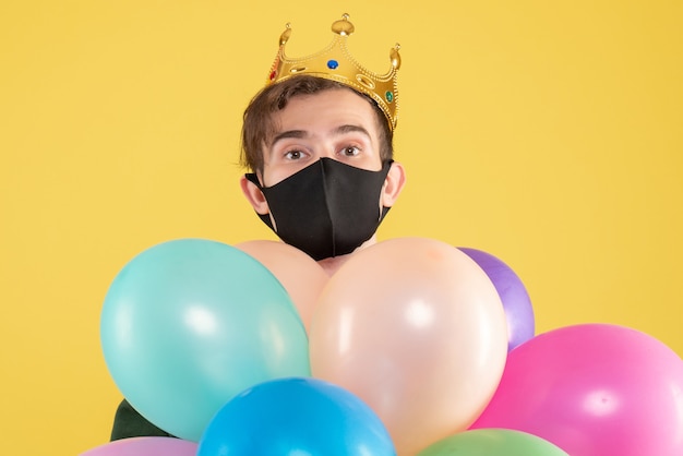 Vorderansicht junger Mann mit Krone und schwarzer Maske, die Luftballons auf Gelb hält