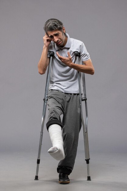 Vorderansicht junger Mann mit Krücken wegen gebrochenem Fuß an der grauen Wand deaktivieren Fuß gebrochenes Unfallbein