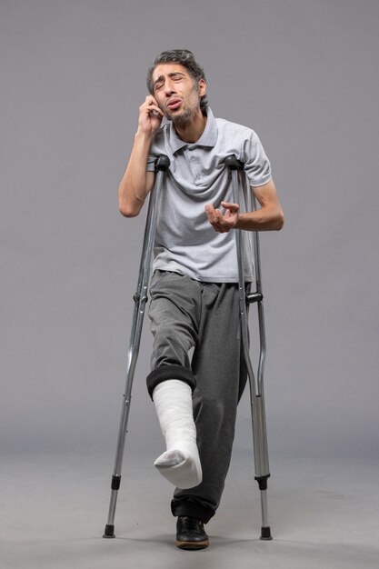 Vorderansicht junger Mann mit Krücken wegen gebrochenem Fuß an grauer Wand deaktivieren Fuß gebrochene Unfallbeine