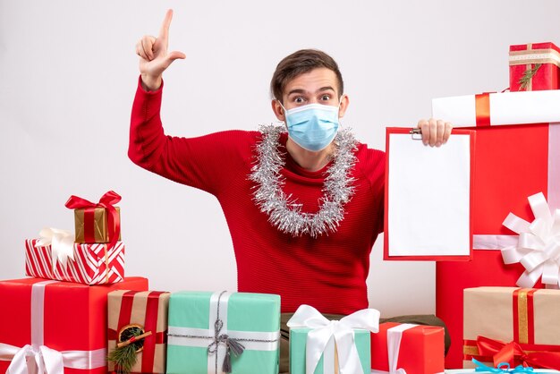 Vorderansicht junger Mann mit mas zeigendem Finger oben sitzend auf Bodenweihnachtsgeschenken