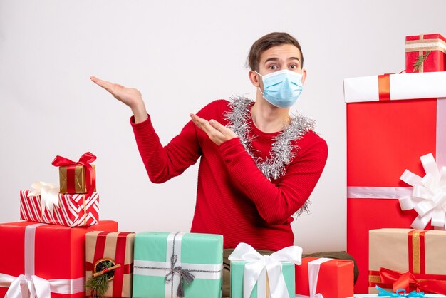 Vorderansicht junger Mann mit Maske, die auf etwas zeigt, das um Weihnachtsgeschenke sitzt