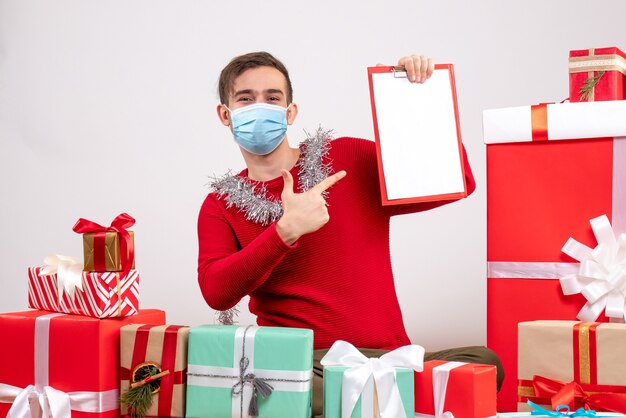 Vorderansicht junger Mann mit Maske, die auf Zwischenablage zeigt, die um Weihnachtsgeschenke sitzt