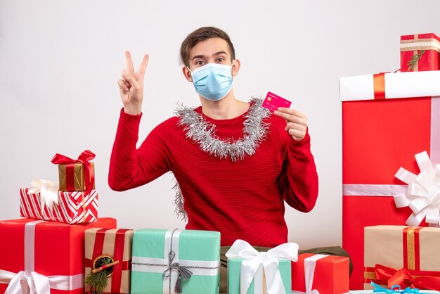 Vorderansicht junger Mann mit Maske, die Siegesfriedenszeichen macht, das um Weihnachtsgeschenke sitzt