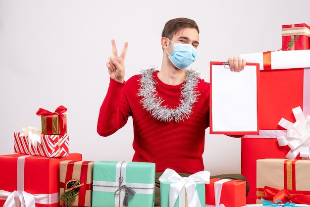 Vorderansicht junger Mann mit Maske, die Siegeszeichen hält, das Klemmbrett hält, das auf Bodenweihnachtsgeschenken sitzt