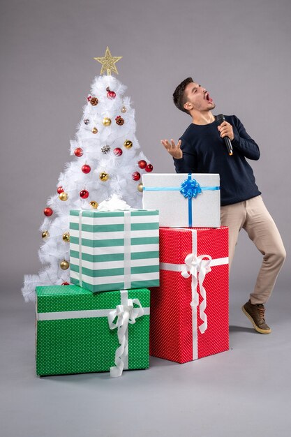 Vorderansicht junger Mann mit Mikrofon mit Geschenken auf grauem Boden Weihnachtsfeiertag neues Jahr