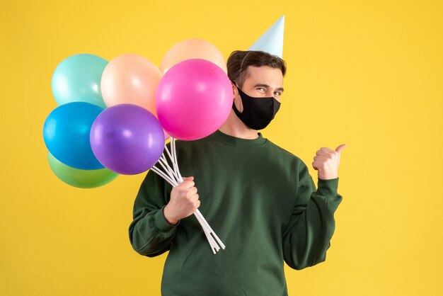 Vorderansicht junger Mann mit Partykappe, die Ballons hält, die auf gelbem Hintergrund stehen