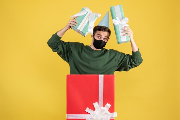 Vorderansicht junger Mann mit Partykappe, die Geschenke hält, die hinter großer Geschenkbox auf gelbem Hintergrund stehen