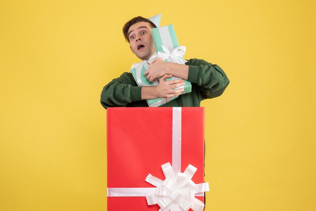 Vorderansicht junger Mann mit Partykappe, die Geschenke hält, die hinter großer Geschenkbox auf gelbem Hintergrund stehen