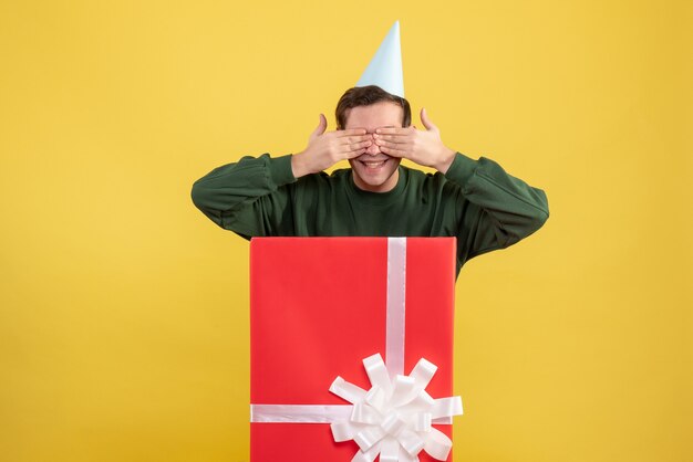 Vorderansicht junger Mann mit Partykappe, die Hände auf seine Augen stellt, die hinter großer Geschenkbox auf gelbem Hintergrund stehen