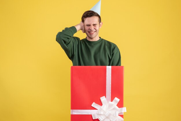 Vorderansicht junger Mann mit Partykappe, die seine Ohren hält, die hinter großer Geschenkbox auf gelbem Hintergrund stehen
