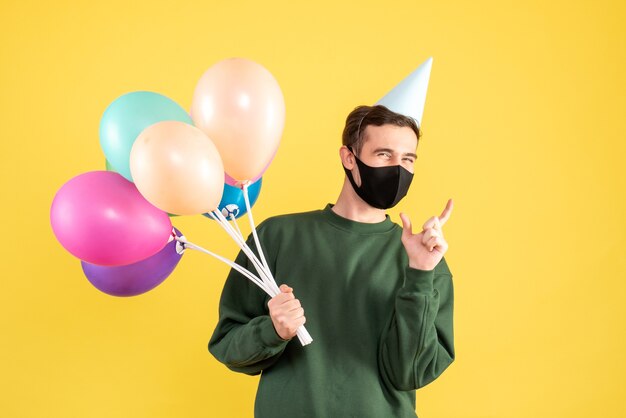 Vorderansicht junger Mann mit Partykappe und bunten Luftballons, die auf gelb stehen