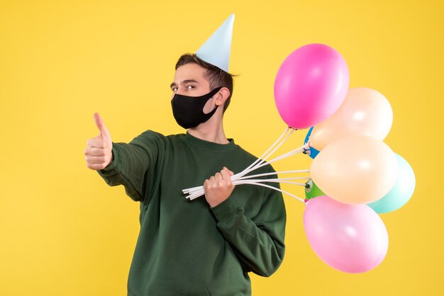 Vorderansicht junger Mann mit Partykappe und bunten Luftballons, die Daumen auf Zeichen stehen, das auf Gelb steht