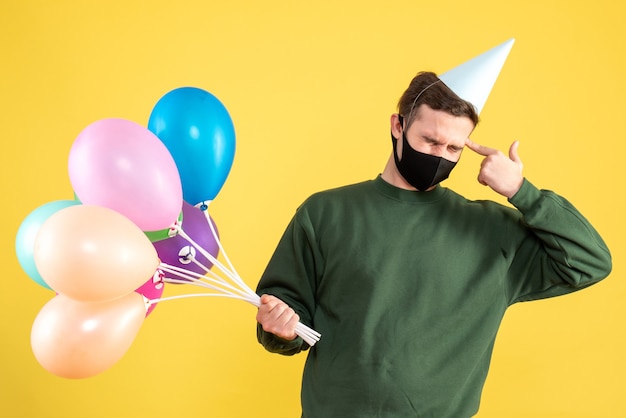 Vorderansicht junger Mann mit Partykappe und bunten Luftballons, die Fingerpistole zu seiner Schläfe stellen, die auf Gelb steht
