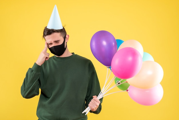 Vorderansicht junger Mann mit Partykappe und bunten Luftballons, die seinen Kopf stehen auf Gelb halten