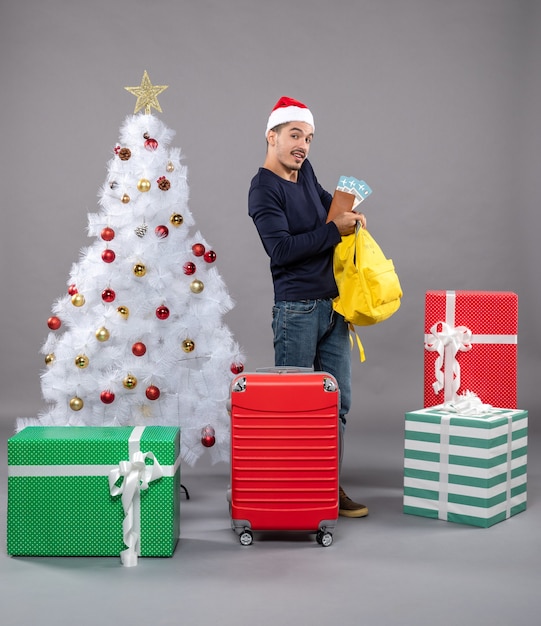 Vorderansicht junger Mann mit Reiseticket und rotem Koffer, der seinen gelben Rucksack um Geschenke auf grauem isoliert überprüft