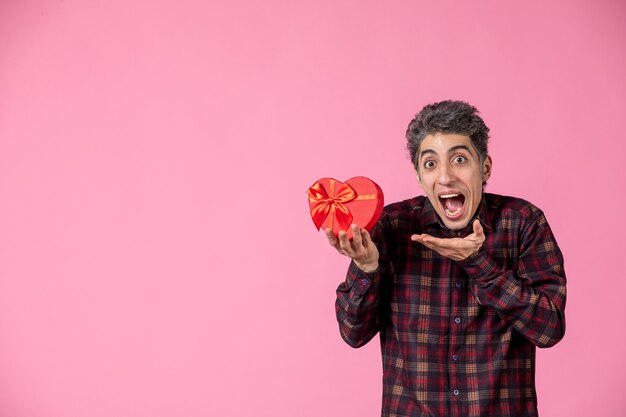 Vorderansicht junger Mann mit rotem herzförmigem Geschenk an rosa Wand