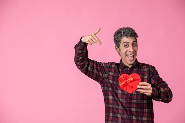 Vorderansicht junger Mann mit rotem herzförmigem Geschenk an rosa Wand