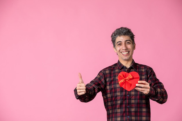 Vorderansicht junger Mann mit rotem herzförmigem Geschenk an rosa Wand