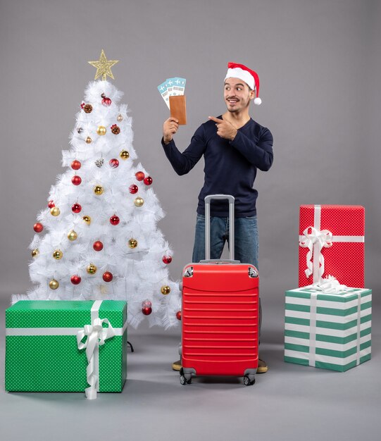 Vorderansicht junger Mann mit rotem Koffer, der seine Reisetickets um weißen Weihnachtsbaum und Geschenke auf Grau zeigt