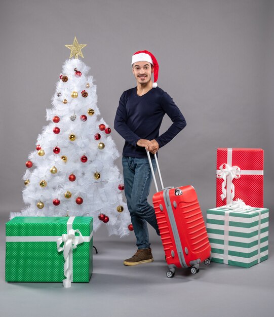 Vorderansicht junger Mann mit roter Reisetasche, der in der Nähe des Weihnachtsbaums auf isoliert steht