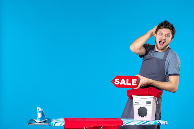 Vorderansicht junger Mann mit roter Verkaufsschrift in den Händen auf blauem Hintergrund Hausarbeit Einkaufen Waschmaschine Bügeleisen Wäsche