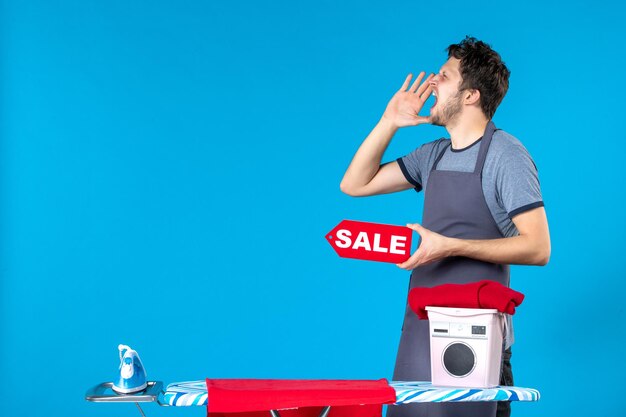 Vorderansicht junger Mann mit roter Verkaufsschrift in den Händen auf blauem Hintergrund Hausarbeit Wäsche Einkaufen Waschmaschine Reinigung Bügeleisen