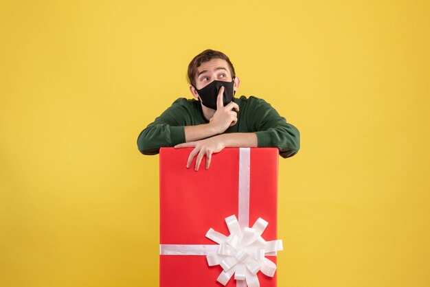 Vorderansicht junger Mann mit schwarzer Maske, die hinter großer Geschenkbox auf Gelb steht