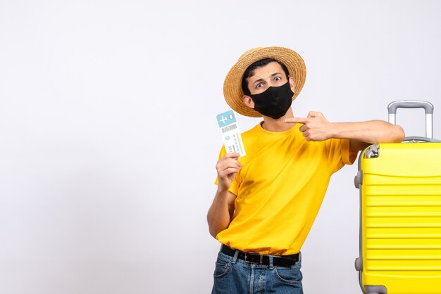 Vorderansicht junger Mann mit Strohhut, der nahe gelbem Koffer steht, der auf Reiseticket zeigt