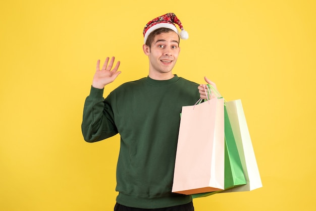 Vorderansicht junger Mann mit Weihnachtsmütze, die Einkaufstaschen hält, die auf gelbem Hintergrund stehen