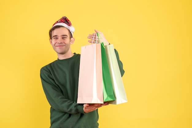 Vorderansicht junger Mann mit Weihnachtsmütze, die Einkaufstaschen hält, die auf gelbem Hintergrund stehen