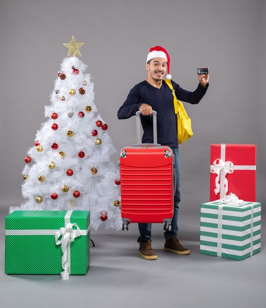Vorderansicht junger Mann mit Weihnachtsmütze mit rotem Koffer, der eine Karte um bunte Geschenke auf grauem isoliert zeigt
