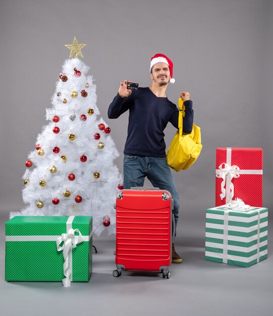 Vorderansicht junger Mann mit Weihnachtsmütze und rotem Koffer, der seinen gelben Rucksack auf grauem isoliert nimmt