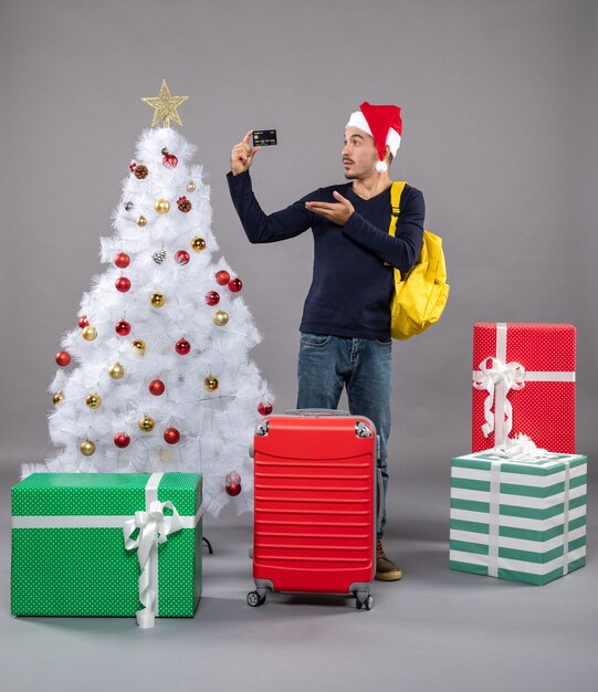 Vorderansicht junger Mann mit Weihnachtsmütze zeigt Karte in der Nähe von Weihnachtsbaum und präsentiert auf grau isoliert