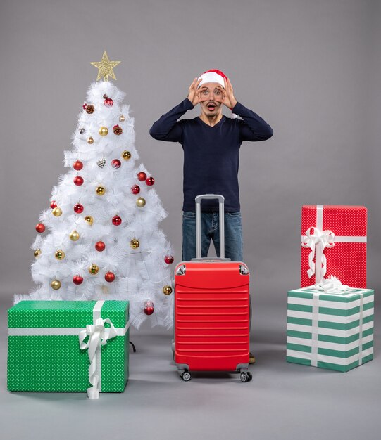 Vorderansicht junger Mann mit weit geöffneten Augen in der Nähe von weißem Weihnachtsbaum und bunten Geschenken auf grau isoliert
