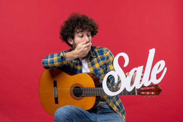 Vorderansicht junger Mann sitzt mit Gitarre auf der roten Wand spielen Konzertmusiker Verkauf Musik Farbapplaus live