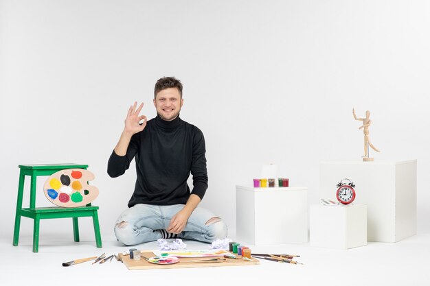 Vorderansicht junger Mann sitzt um Farben und Quasten zum Zeichnen auf der weißen Wand Farbe Bild malen Kunst Malerei Künstler zeichnen