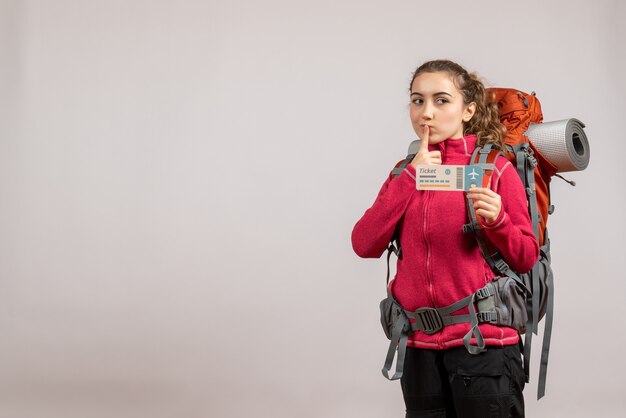 Vorderansicht junger Reisender mit großem Rucksack, der das Reiseticket hochhält und den Finger auf den Mund legt