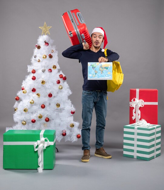 Vorderansicht junger weihnachtsmann mit gelbem rucksack, der karte in der nähe des weihnachtsbaums hält und präsentiert auf grauem isoliert