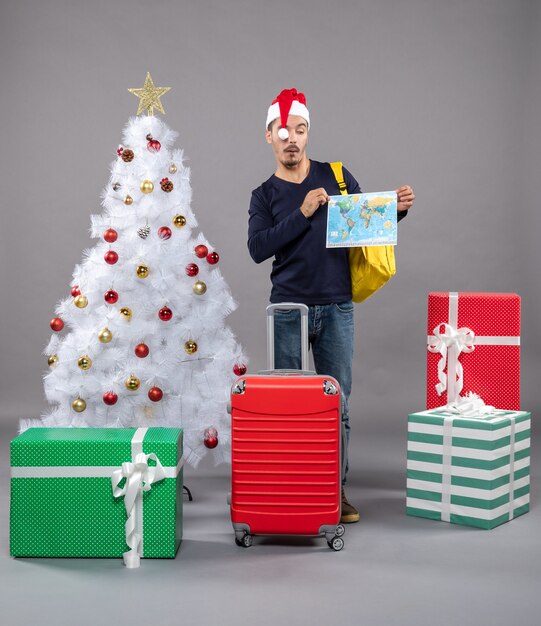 Vorderansicht junger Weihnachtsmann mit rotem Koffer, der Karte mit beiden Händen auf grauem isoliert hält