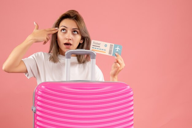 Vorderansicht junges Mädchen mit rosa Koffer mit Ticket, das Fingerpistole auf ihre Schläfe legt