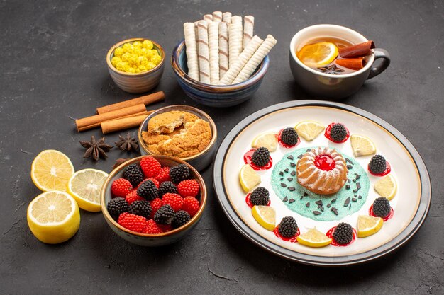 Vorderansicht kleiner leckerer Kuchen mit Zitronenscheiben Bonbons und Tasse Tee auf dunklem Hintergrund Kekskuchen Obst Zitrus süße Kekse