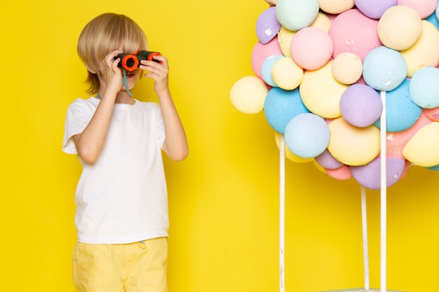 Vorderansicht kleiner süßer Junge im weißen T-Shirt zusammen mit bunten Luftballons auf gelbem Schreibtisch