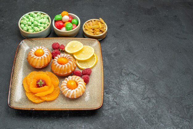 Vorderansicht köstliche kleine Kuchen mit Zitronenscheiben Mandarinen und Bonbons auf dunklem Schreibtisch Tee Fruchtkeks süße Kekskuchen