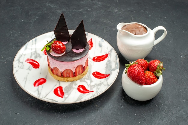 Vorderansicht köstlicher käsekuchen mit erdbeer- und schokoladenschüssel mit erdbeeren und schokolade auf ovalem teller auf dunklem hintergrund