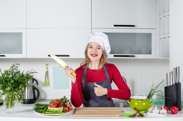 Vorderansicht lächelnde junge Frau in Schürze, die das Messer hochhält, das Daumen hochgibt